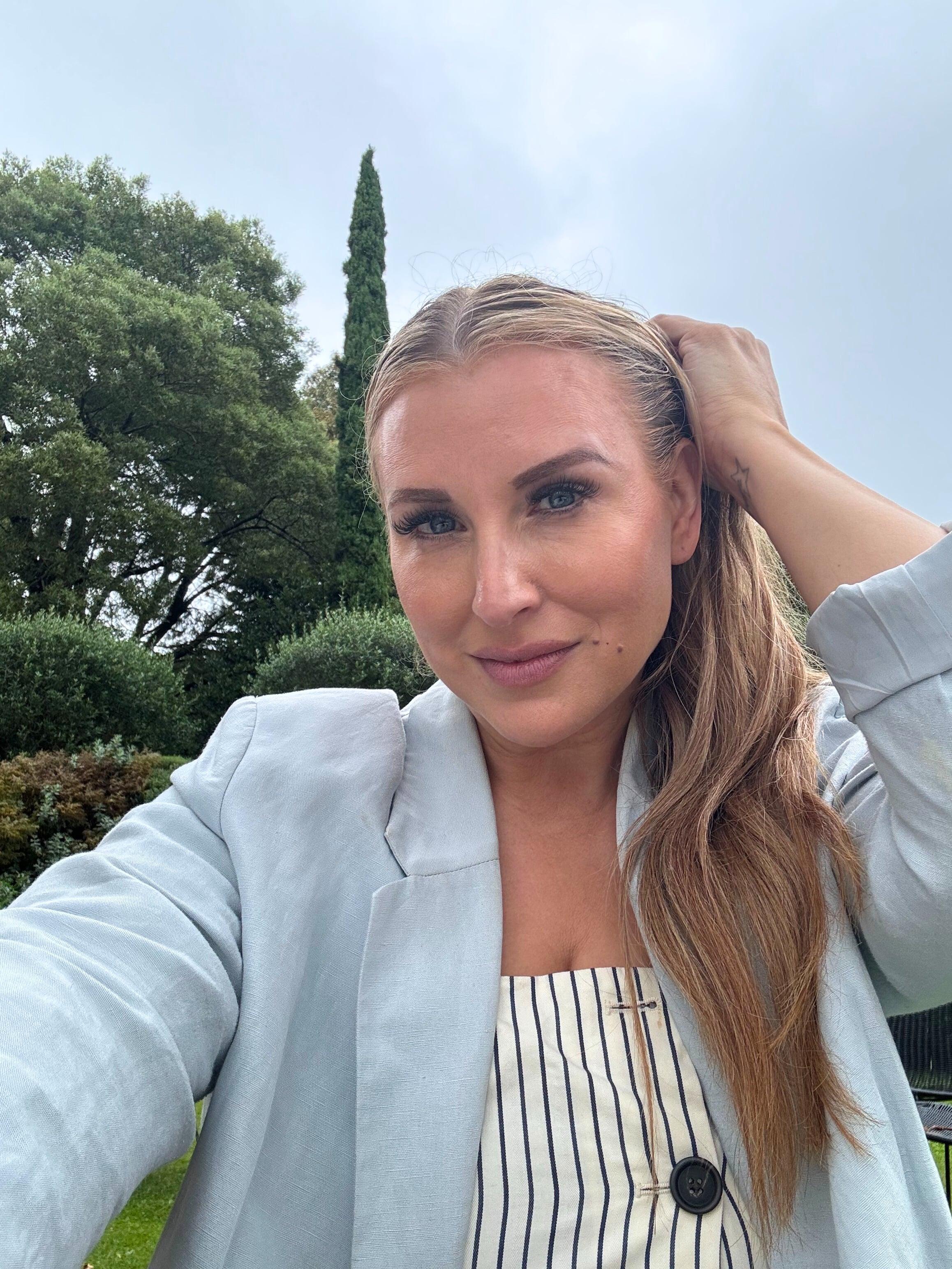 Quinn in a light blue blazer and striped top in front of a group of trees