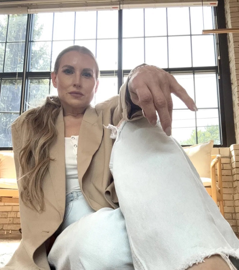 Quinn in white jeans and a tan over coat, backlit by warehouse windows, sitting down on the floor and looking at the camera