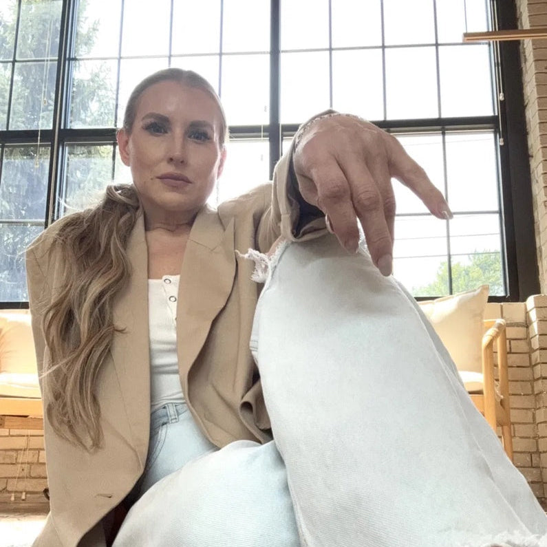 Quinn in white jeans and a tan over coat, backlit by warehouse windows, sitting down on the floor and looking at the camera