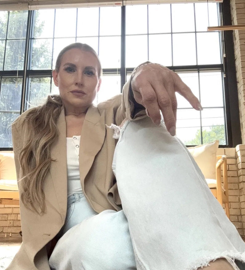 Quinn in white jeans and a tan over coat, backlit by warehouse windows, sitting down on the floor and looking at the camera
