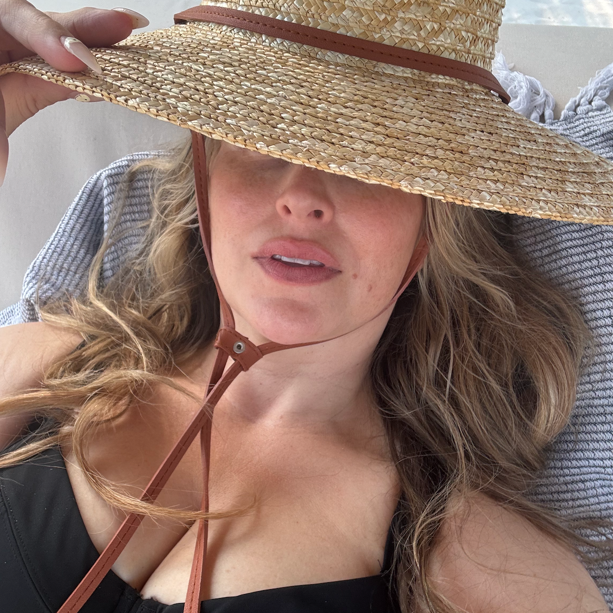 A selfie of Quinn in a black bathing suit laying on a towel and a straw hat obscuring her face