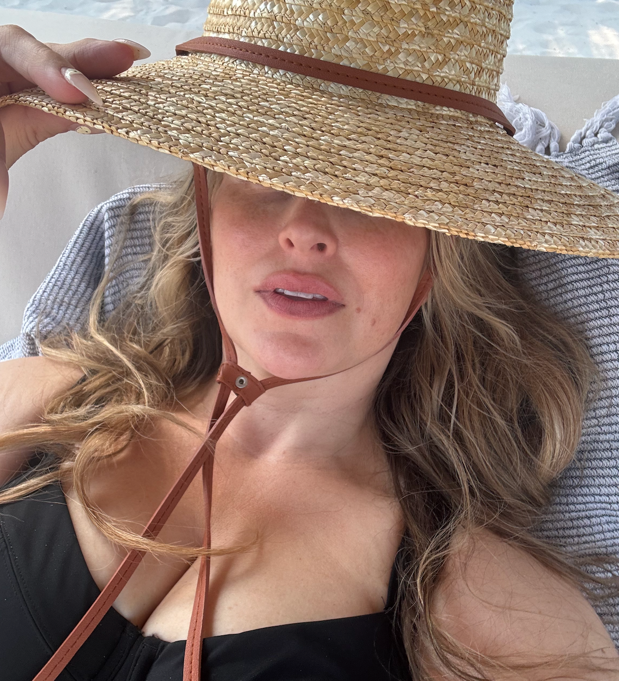 A selfie of Quinn in a black bathing suit laying on a towel and a straw hat obscuring her face