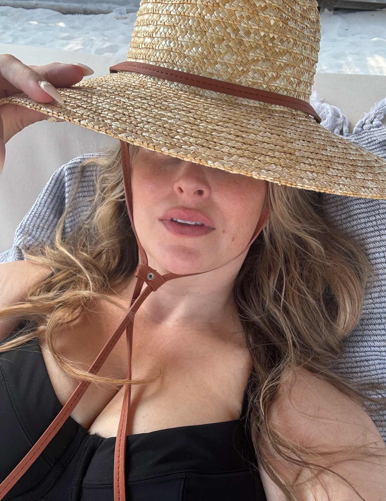A selfie of Quinn in a black bathing suit laying on a towel and a straw hat obscuring her face