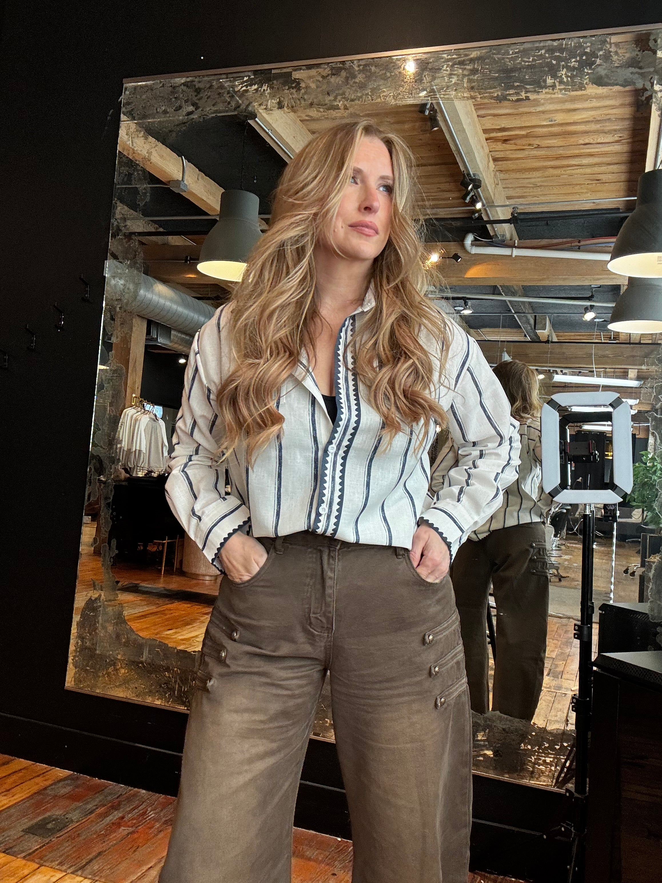 A woman with long wavy blonde hair stands in front of a large mirror in a retail store, wearing a white button-down shirt with thin black vertical stripes and brown wide-leg pants.